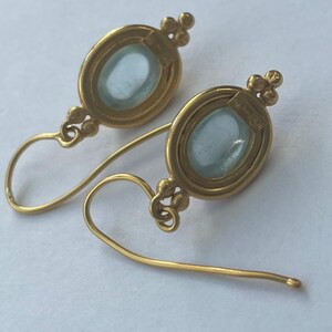 May include: A pair of gold-colored earrings with oval, light blue gemstones. The earrings have a delicate design with a small cluster of beads at the top and a curved hook. The gemstones are set in a gold frame.
