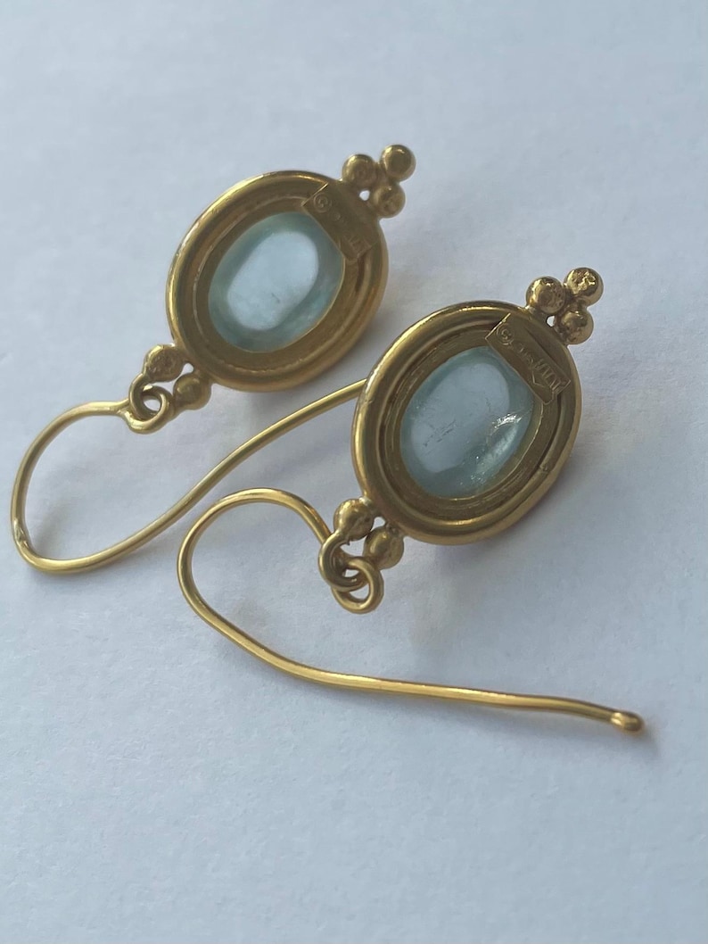 May include: A pair of gold-colored earrings with oval, light blue gemstones. The earrings have a delicate design with a small cluster of beads at the top and a curved hook. The gemstones are set in a gold frame.