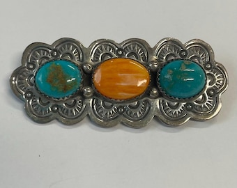 Può includere: Un fermaglio d'argento con tre pietre: turchese, arancio e turchese. Il fermaglio ha un bordo decorativo a festoni.