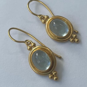 May include: A pair of gold-colored earrings with oval, light blue gemstones. The earrings have a hook closure and are accented with small, round gold beads. The gemstones are set in a gold bezel.