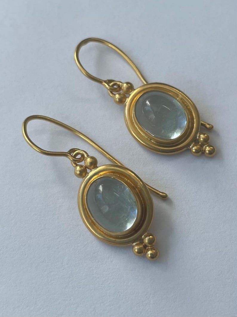 May include: A pair of gold-colored earrings with oval, light blue gemstones. The earrings have a hook closure and are accented with small, round gold beads. The gemstones are set in a gold bezel.