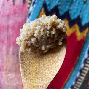 May include: A close-up of a wooden spoon filled with a light brown, grainy substance. The spoon is held against a colourful, patterned background.
