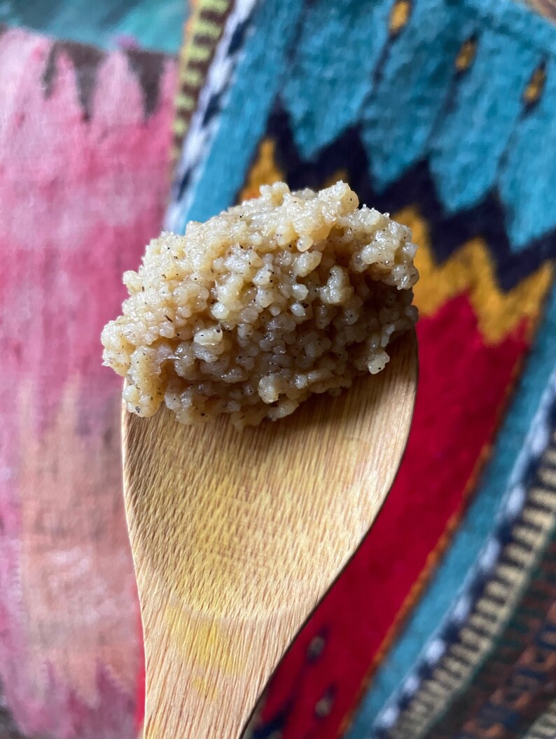 May include: A close-up of a wooden spoon filled with a light brown, grainy substance. The spoon is held against a colourful, patterned background.