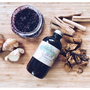 May include: A wooden cutting board with various ingredients for making a homemade elderberry elixir, including a jar of elderberry jam, a bottle of elderberry syrup labeled "elderberry elixir wellness syrup", ginger root, cinnamon sticks, and pieces of chaga mushroom.