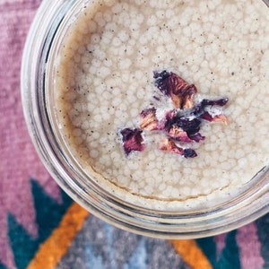 May include: A glass jar filled with a light beige cream with a speckled texture. Dried rose petals are floating on top of the cream.