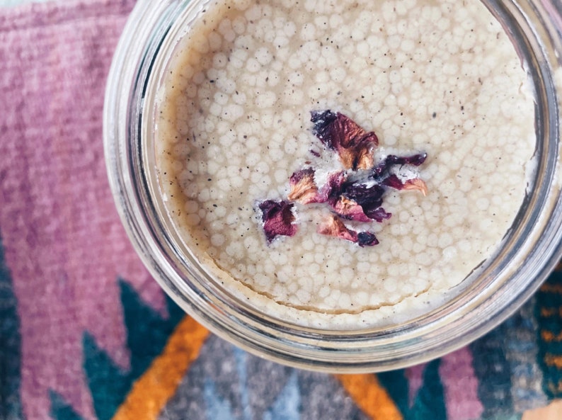 May include: A glass jar filled with a light beige cream with a speckled texture. Dried rose petals are floating on top of the cream.