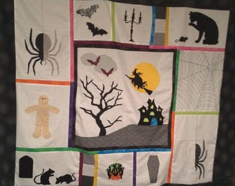 Könnte beinhalten: Eine weiße Steppdecke mit Halloween-Motiv. Die Steppdecke besteht aus verschiedenen Quadraten mit verschiedenen Halloween-Bildern, darunter eine Spinne, eine Hexe, ein Spukhaus, eine Fledermaus, eine Mumie, ein Kessel, ein Spinnennetz und ein Friedhof. Die Steppdecke ist mit einem schwarzen Rand eingefasst.
