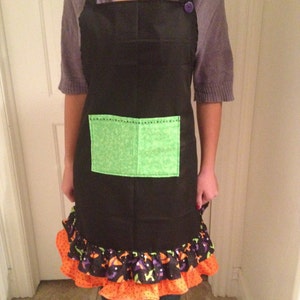 May include: Black apron with a green pocket and a ruffle trim featuring a Halloween theme. The ruffle is orange, black, and purple with a white polka dot pattern.