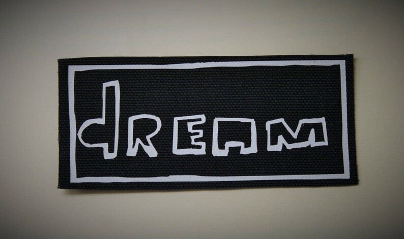 K&ouml;nnte beinhalten: Schwarzer rechteckiger Aufn&auml;her mit wei&szlig;em Rand. Das Wort "DREAM" steht in wei&szlig;en Blockbuchstaben &uuml;ber die Mitte des Aufn&auml;hers geschrieben. Der Aufn&auml;her hat ein strukturiertes Aussehen.