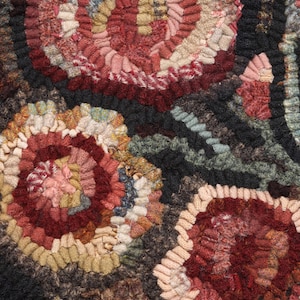 May include: Close-up of a handmade rug with a floral design. The rug features a variety of colors, including red, pink, beige, and green, arranged in a circular pattern. The background is a dark brown color.