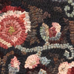 May include: Close-up of a handmade rug with a floral pattern. The rug features a dark background with colorful, textured flowers in shades of red, pink, and beige. The design is intricate and detailed, showcasing the craftsmanship.