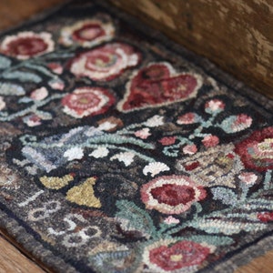 May include: A rectangular, hand-hooked rug with a dark background and a floral design. The rug features red, pink, and white flowers, green leaves, and a red heart. The edges are dark gray, and the rug has the numbers "1818" on it.