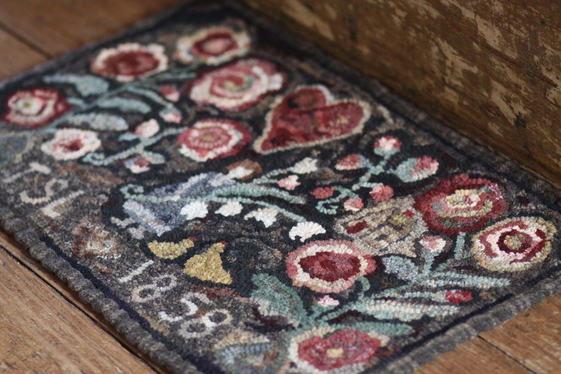 May include: A rectangular, hand-hooked rug with a dark background and a floral design. The rug features red, pink, and white flowers, green leaves, and a red heart. The edges are dark gray, and the rug has the numbers "1818" on it.