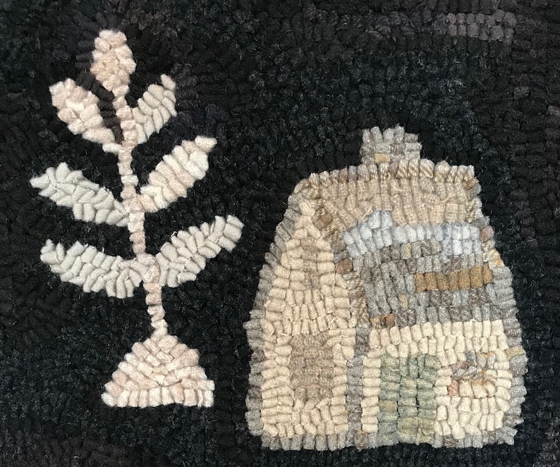 May include: A handmade rug with a black background features a stylized house and a leafy plant. The house is beige, gray, and green, while the plant is beige and white. The rug's texture is visible, adding depth to the design.