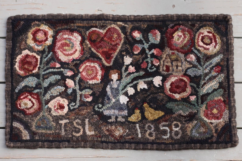 May include: Hand-hooked rug with a dark brown background, featuring a heart, flowers, and a figure. The rug includes the text "TSL 1858" in white. The flowers are in shades of red, pink, and cream, with green leaves.