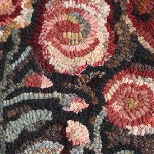 May include: Close-up of a handmade rug with a floral design. The rug features large, stylized flowers in shades of red, pink, and cream against a dark background. The texture is created with looped yarn, giving a three-dimensional effect.