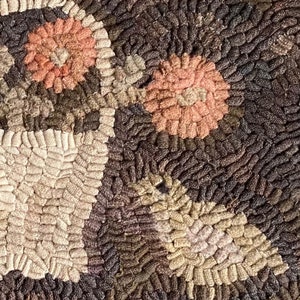 May include: Close-up of a handmade rug with a floral design. The rug features a basket with two pink flowers and green leaves. The background is a mix of brown, beige, and gray tones, creating a textured, rustic appearance.