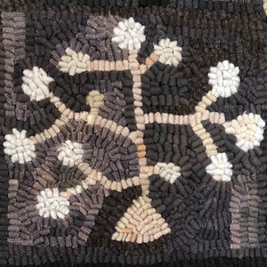 May include: Close-up of a hand-hooked rug with a floral design. The rug features a dark background with a central motif of a stylized tree or plant in shades of cream, gray, and black. The design is symmetrical and textured, with a handmade appearance.