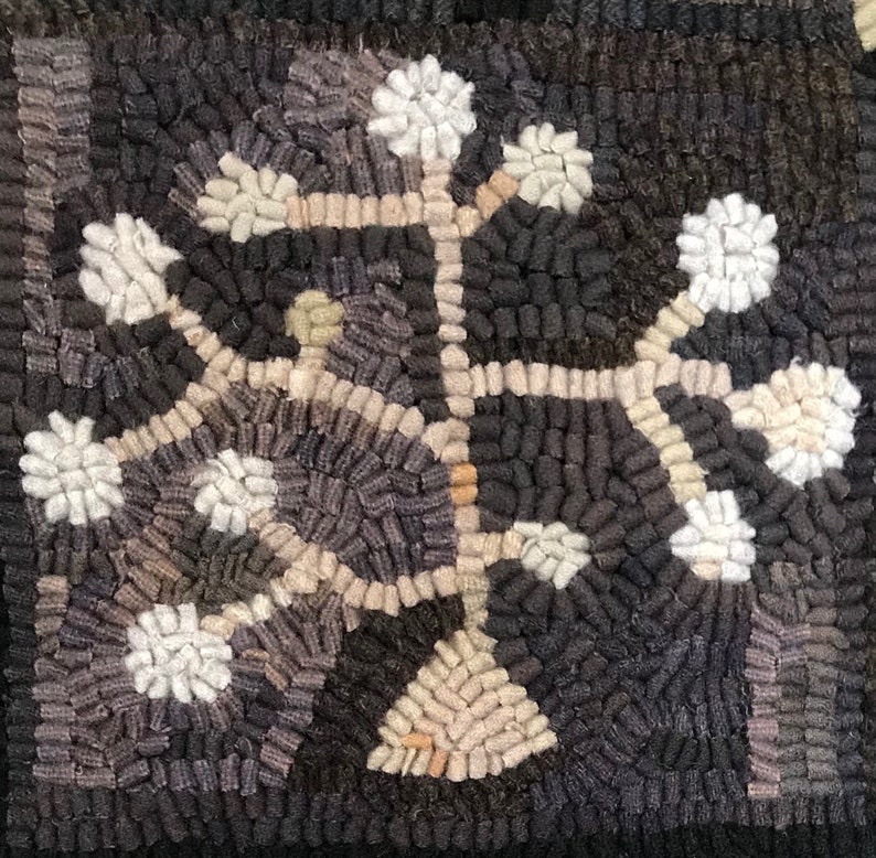 May include: Close-up of a hand-hooked rug with a floral design. The rug features a dark background with a central motif of a stylized tree or plant in shades of cream, gray, and black. The design is symmetrical and textured, with a handmade appearance.