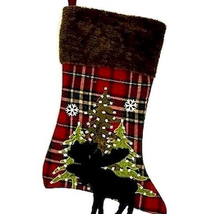 Peut inclure: Chaussette de Noël à motif écossais rouge et noir. Le haut est en fausse fourrure marron. La chaussette présente une silhouette d'orignal noire, des sapins verts et des flocons de neige blancs.