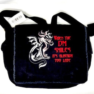 Puede incluir: Bolso mensajero negro con un diseño de dragón blanco y plateado y el texto rojo "When the DM Smiles It's Already Too Late". El bolso tiene una correa para el hombro y una etiqueta adjunta.