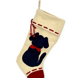 May include: A beige Christmas stocking featuring a black dog silhouette wearing a red bow tie. The stocking has a red heel and trim, with red stitching and a red ribbon with two small bells. A paper clip is attached to the top.