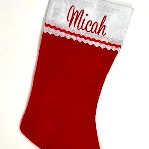 May include: A red Christmas stocking with a white cuff and a red loop for hanging. The name "Micah" is embroidered in red script on the white cuff, which is trimmed with a scalloped red border.
