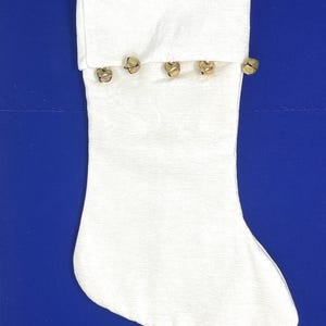 May include: A white Christmas stocking with a textured finish, complete with a hanging loop. The stocking is decorated with five small, gold-coloured bells along the top edge, set against a blue backdrop.
