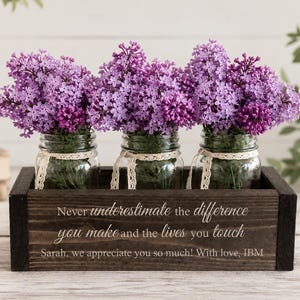 May include: A dark wooden box holding three glass jars, each filled with purple lilacs. The jars are adorned with lace and sit in a dark brown wooden box. The box has the text: "Never underestimate the difference you make and the lives you touch."