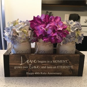 May include: A wooden box with three mason jars filled with purple and blue hydrangeas. The box has a white inscription that reads "Love begins in a MOMENT, grows over time, and lasts an ETERNITY. Happy 40th Ruby Anniversary."