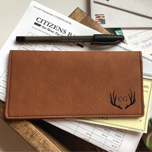 Puede incluir: Una funda rectangular para chequera de color canela con superficie texturizada y costuras negras en los bordes. El frente presenta un dise&ntilde;o grabado con l&aacute;ser en negro de dos astas de ciervo que enmarcan las iniciales CG en fuente serif.