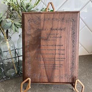 May include: A wooden memorial plaque with the text "Maria's Bookshelf" and an inscription honoring Maria A. Walbrandt, including her birth and death years, and other details. The plaque is displayed on a gold-colored stand.