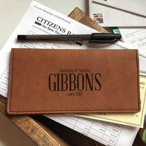 May include: A rectangular tan faux leather checkbook cover with black stitching. The front is engraved with the text "Jonathan & Sandra GIBBONS since 2013". The cover is positioned on top of bank documents and a black pen, with a small succulent visible.