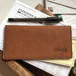 May include: A rectangular tan checkbook cover with a textured surface and black stitching along the edges. The name Audrey is engraved in a black cursive script on the bottom right corner of the cover. The cover is placed over bank documents.