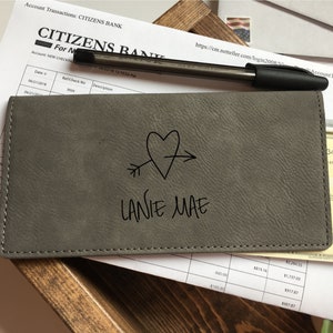 May include: A rectangular gray checkbook cover with a textured finish, featuring a black line drawing of a heart pierced by an arrow and the name LANIE MAE printed in black text. The cover is stitched along the edges with matching gray thread.