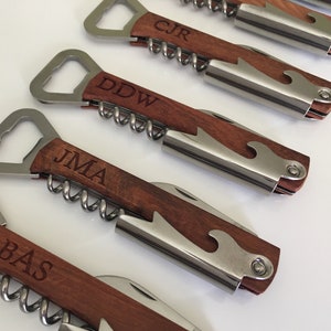 May include: Five personalized bottle openers with wooden handles. Each opener has a corkscrew, bottle opener, and a small knife. The handles are engraved with the initials "BAS", "JMA", "DDW", "CJR", and a fifth opener with an unreadable inscription.