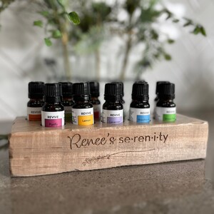 May include: A wooden essential oil holder with the inscription "Renee's serenity" and a row of ten small glass bottles with black caps and colourful labels. The labels include the words "REVIVE", "Purity", "Lemon", "Lavender", and "Breathe".