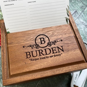 May include: A wooden recipe box with a personalized inscription that reads "From the home of Burden, Recipes loved by our family". The box has a slot for recipe cards and a lid that lifts to reveal the interior.