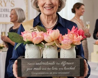 Puede incluir: Una caja rectangular de madera oscura que contiene tres frascos de vidrio blanco llenos de peonías artificiales de color rosa y melocotón. La caja presenta el texto grabado: Never underestimate the difference you made and the lives you touched. Congratulations on your retirement!