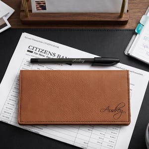 May include: A tan rectangular checkbook cover with a textured surface and the name Audrey embossed in black script. A black pen with the name Audrey printed on the barrel rests on top of the cover, which is placed on a Citizens Bank document.