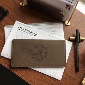 以下が含まれることがあります： 中央に文字「R」が刻印された円形のリースデザインがレーザー彫刻された、長方形のトープ色の小切手帳カバー。カバーの縁には目に見えるステッチが施されており、Citizens Bankの小切手登録簿の上に置かれています。