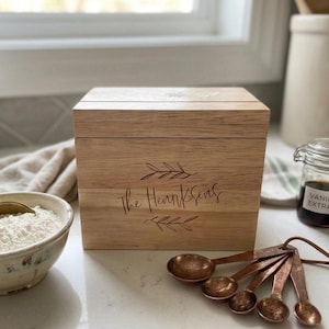 May include: A wooden recipe box with the name "The Hennksens" engraved on the front, accompanied by copper measuring spoons and a bowl of flour. A small jar of vanilla extract and a white ceramic jar are also visible.
