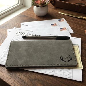 May include: A rectangular gray checkbook cover with a matte, suede-like texture. The front features a laser-engraved design of deer antlers flanking the initials AB. The cover is stitched along the edges with matching gray thread.