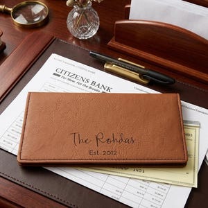 May include: A rectangular tan leather checkbook cover featuring the embossed text The Rohdas and Est. 2012. The cover is placed on top of a Citizens Bank document, with a black pen, a wooden letter holder, and a magnifying glass nearby on a desk.