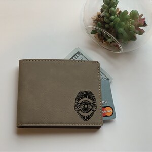 May include: A tan bi-fold wallet featuring a black engraved police badge emblem on the front. The wallet contains a one hundred dollar bill and a grey credit card with a visible Mastercard logo tucked into the interior slots.