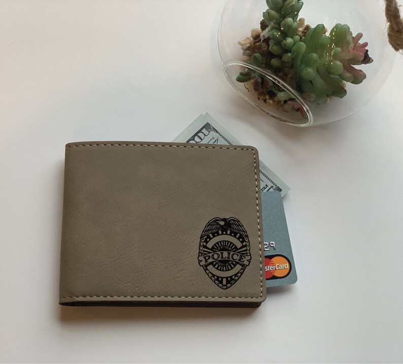 May include: A tan bi-fold wallet featuring a black engraved police badge emblem on the front. The wallet contains a one hundred dollar bill and a grey credit card with a visible Mastercard logo tucked into the interior slots.