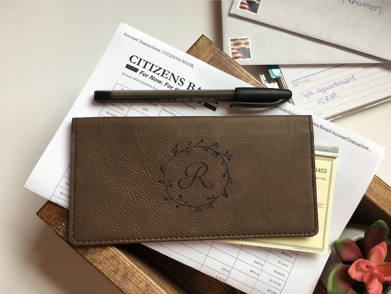 May include: A brown rectangular checkbook cover with a textured surface featuring a dark engraved circular wreath design surrounding the letter R. The cover is positioned on top of white financial documents and a black pen, with visible text Citizens Bank.