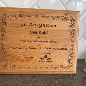 May include: A rectangular wooden plaque with engraved text and floral border. The text reads "In Recognition Bea Kohl for 2500 Hours of Volunteer Service to Wood County Master Gardener Association May 2026". The plaque has a natural wood grain finish.