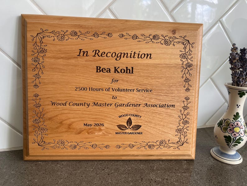 May include: A rectangular wooden plaque with engraved text and floral border. The text reads "In Recognition Bea Kohl for 2500 Hours of Volunteer Service to Wood County Master Gardener Association May 2026". The plaque has a natural wood grain finish.