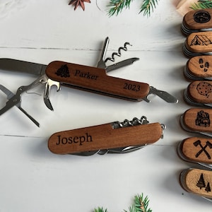 May include: Wooden multi-tool pocket knives with various tools, including a blade, corkscrew, and bottle opener. One knife is open, displaying the tools. The knives are engraved with names and small icons, such as a tree or a tent.